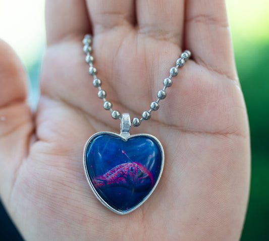 Painting of a neon pink and purple anglerfish in a steel heart pendant with a ball chain necklace against a hand
