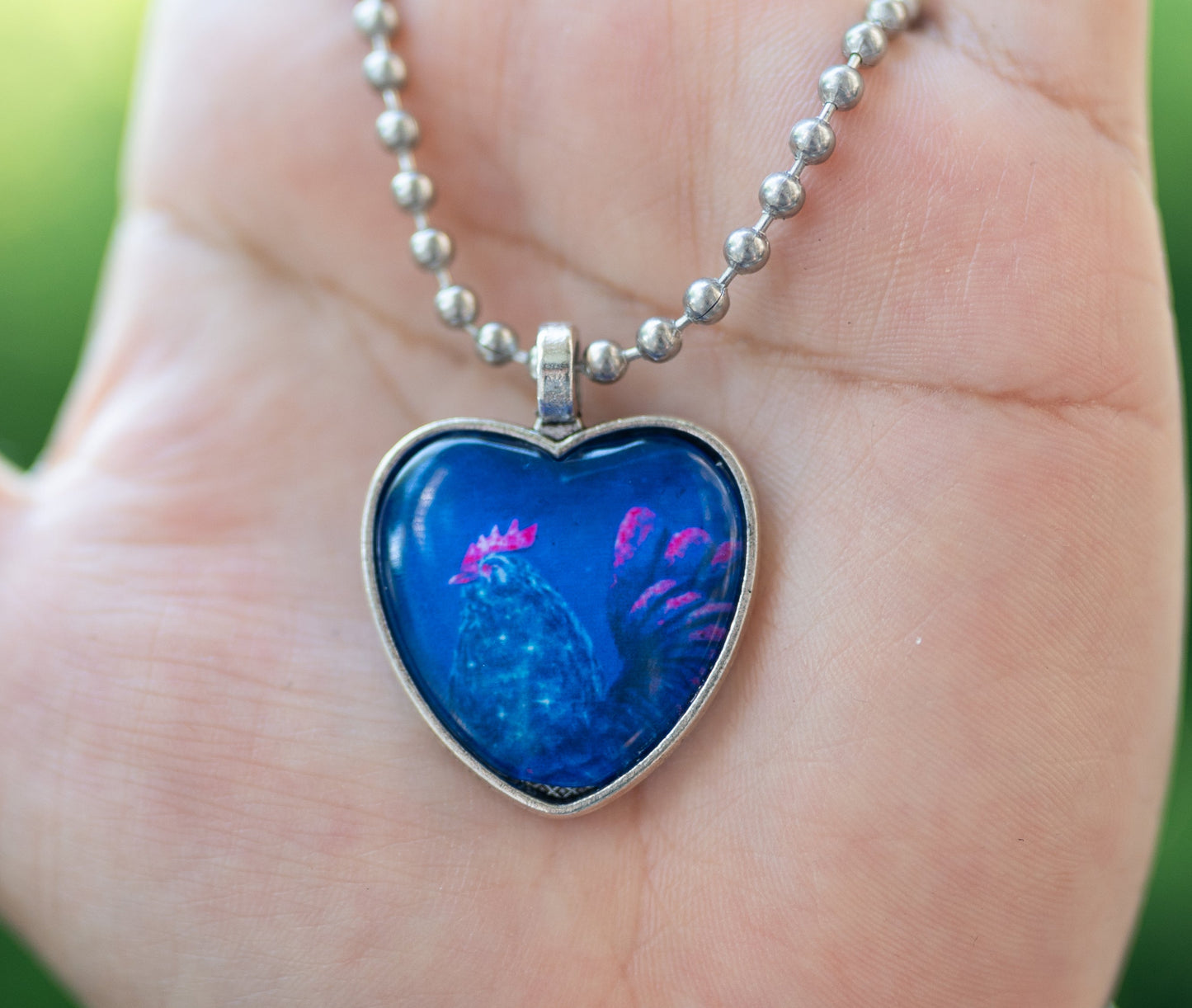 Blue heart-shaped pendant with a blue and pink chicken on a silver chain held in a hand with a blurred green background