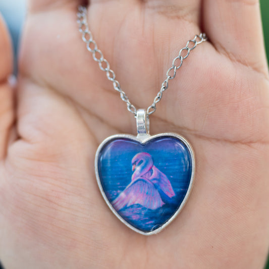 Heart-shaped pendant with a blue and pink design of a swan in water held in a hand against a blurred green background