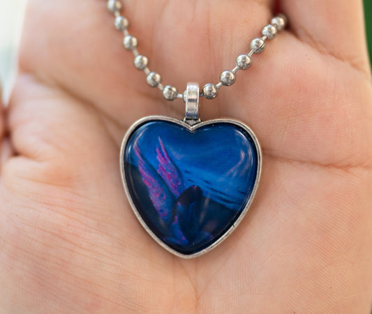 Heart-shaped blue pendant  of an angel with pink wings on a silver chain held in a hand with a blurred green background