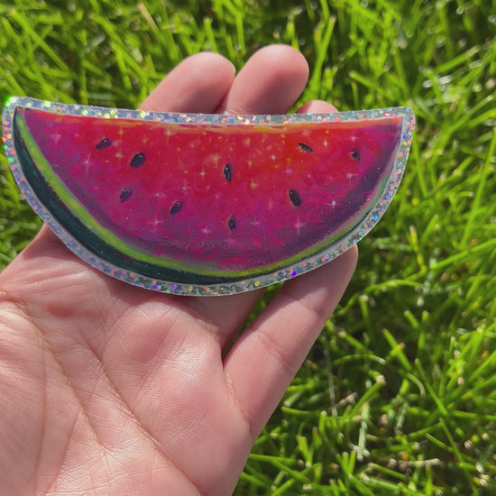Video of a bright neon pink, red, and purple watermelon painting with seeds. Outlined with glitter. The sticker is moved back and forth to show the glitter effect. 